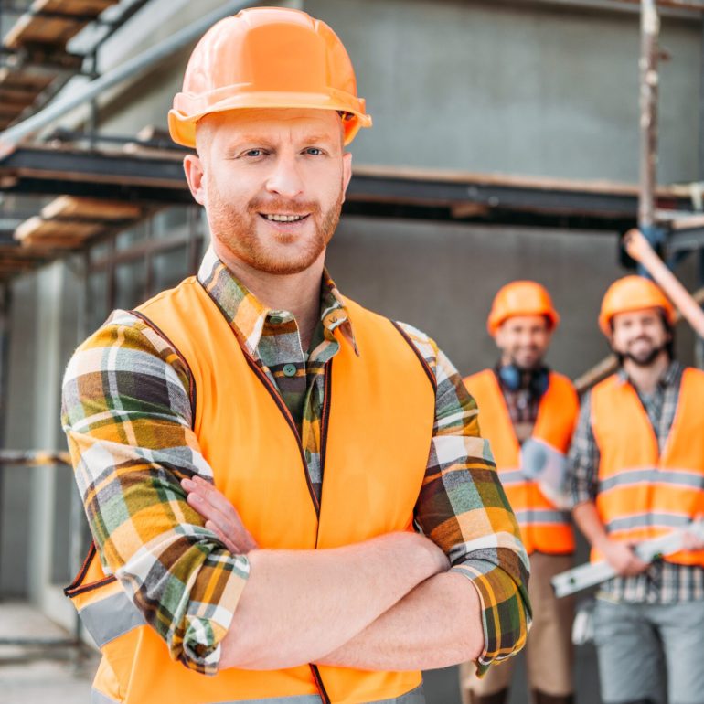 smiling builder standing at construction site with crossed arms while his colleagues standing